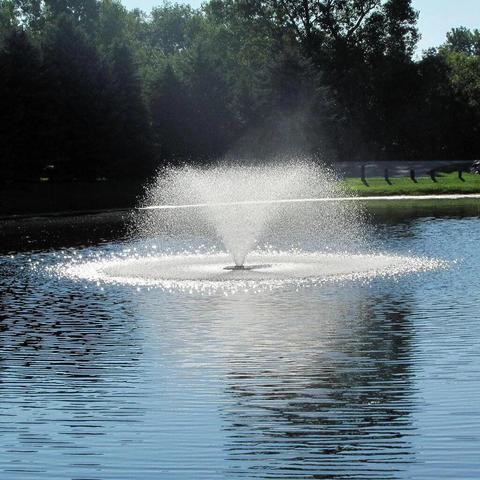 Water spraying from decorative pond fountains in a calm outdoor pond surrounded by trees.