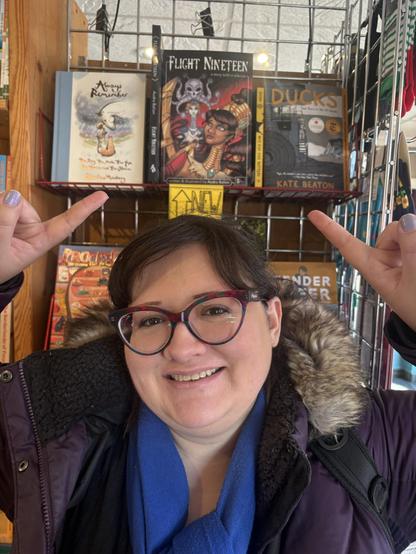 Photo of artist Audra Balion smiling and pointing above her head at her Flight Nineteen sitting upon a bookshelf.
