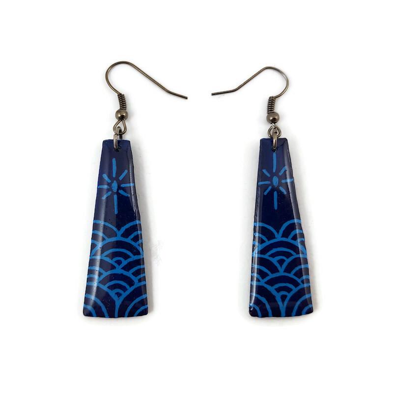 Boucles d'oreilles fantaisie pendantes en forme de longs trapèzes bleus marine ornés de vagues et d'un soleil stylisés bleus ciel.
Les perles sont réalisées en CD recyclé, découpées et peintes à la main, puis recouvertes de résine époxy.
Les crochets sont en métal argenté, mais sont disponibles dans d'autres coloris et métaux.