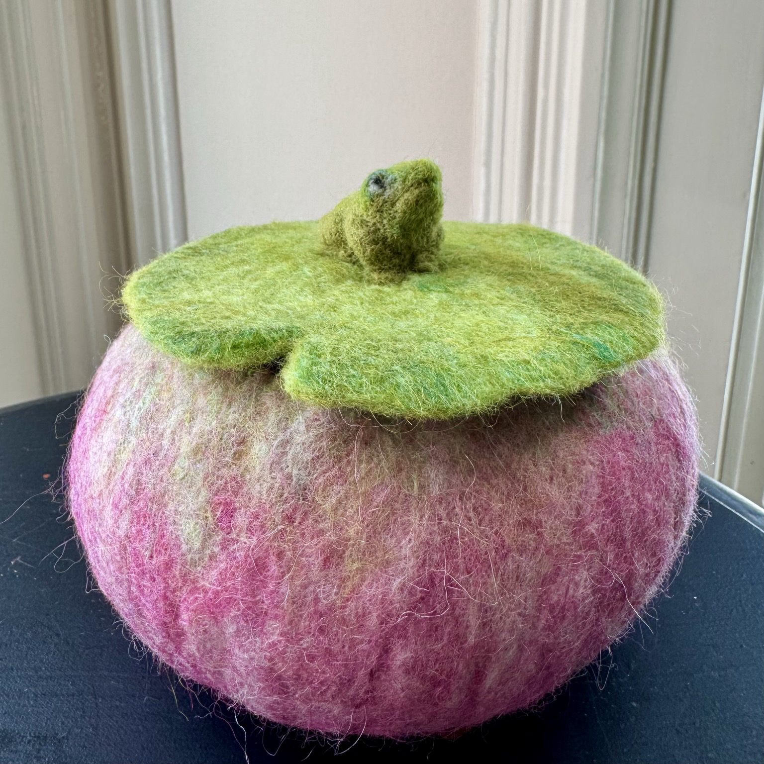 A green needle felted frog sits atop a pink wet felted vessel and its lily pad lid.