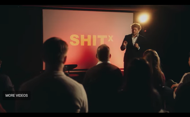 A person with a bright light next to his head stands next to a projection screen with the word SHIT [superscript]X[/superscript] in yellow text on a red background with an audience of onlookers.