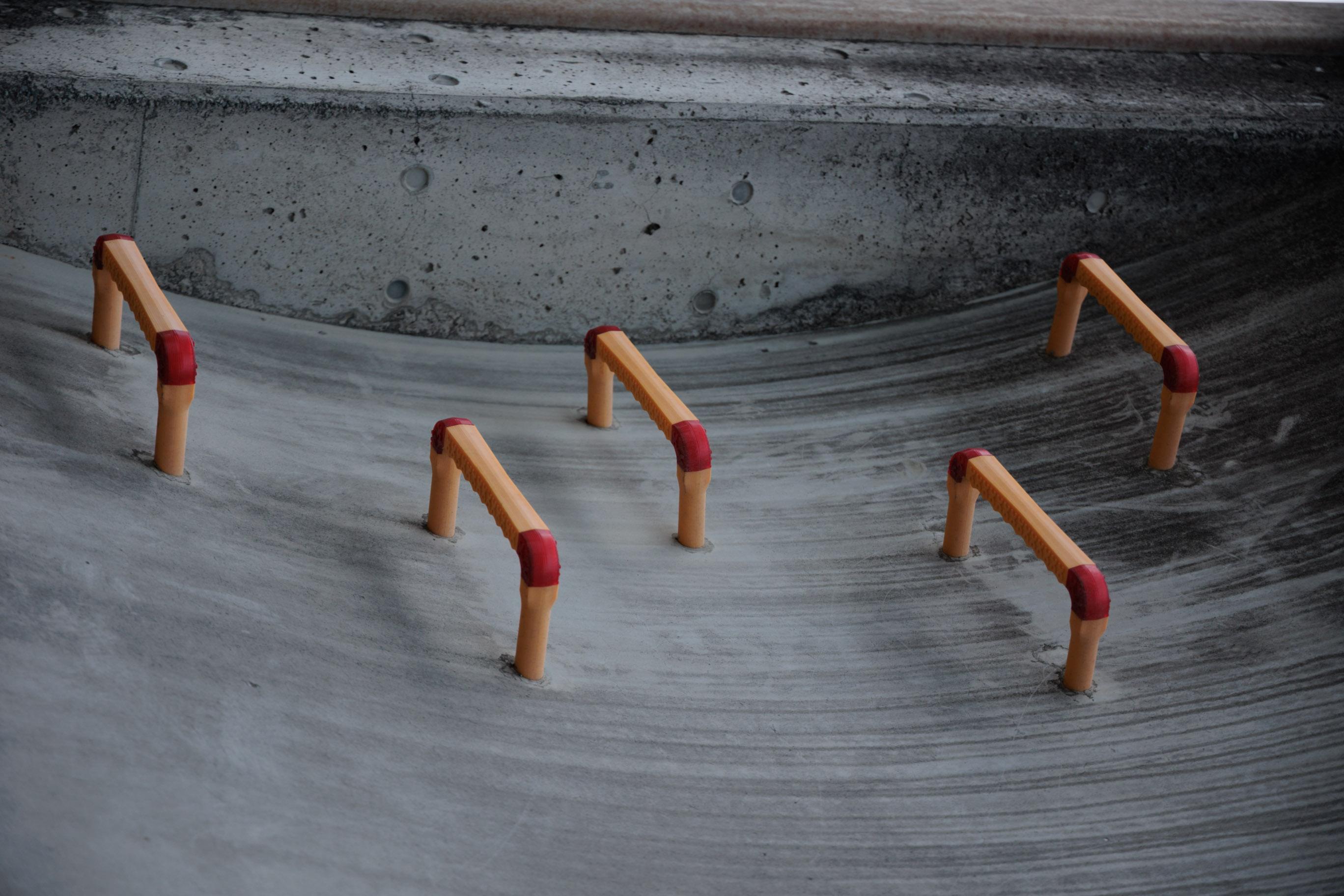 Five small, curved metal rails are positioned on a smooth, gray concrete surface. Each rail features an orange base with red ends.