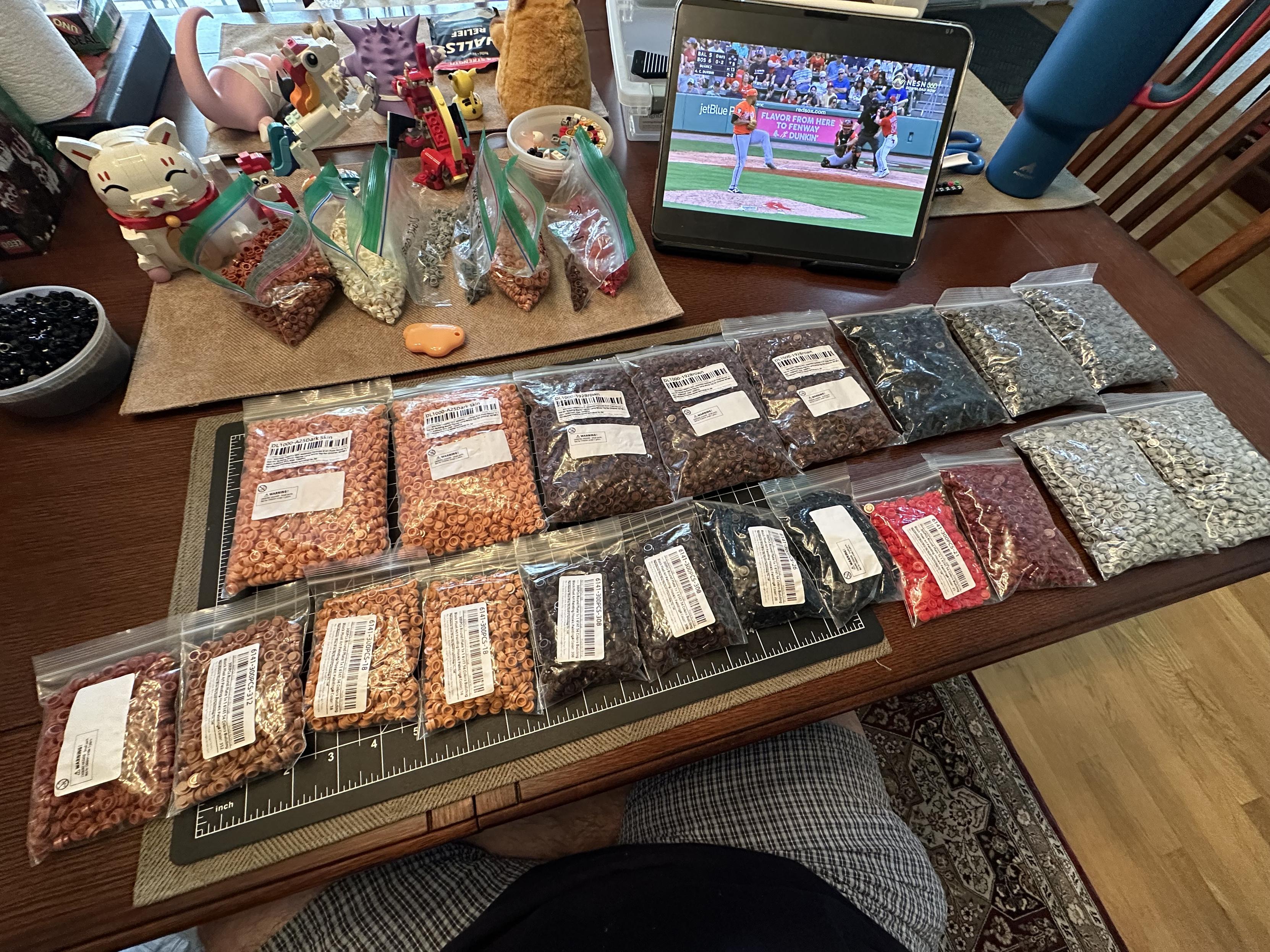 My dining room table covered in many large bags of single stud LEGO pieces of various colors. A baseball game is playing on an iPad in the background.