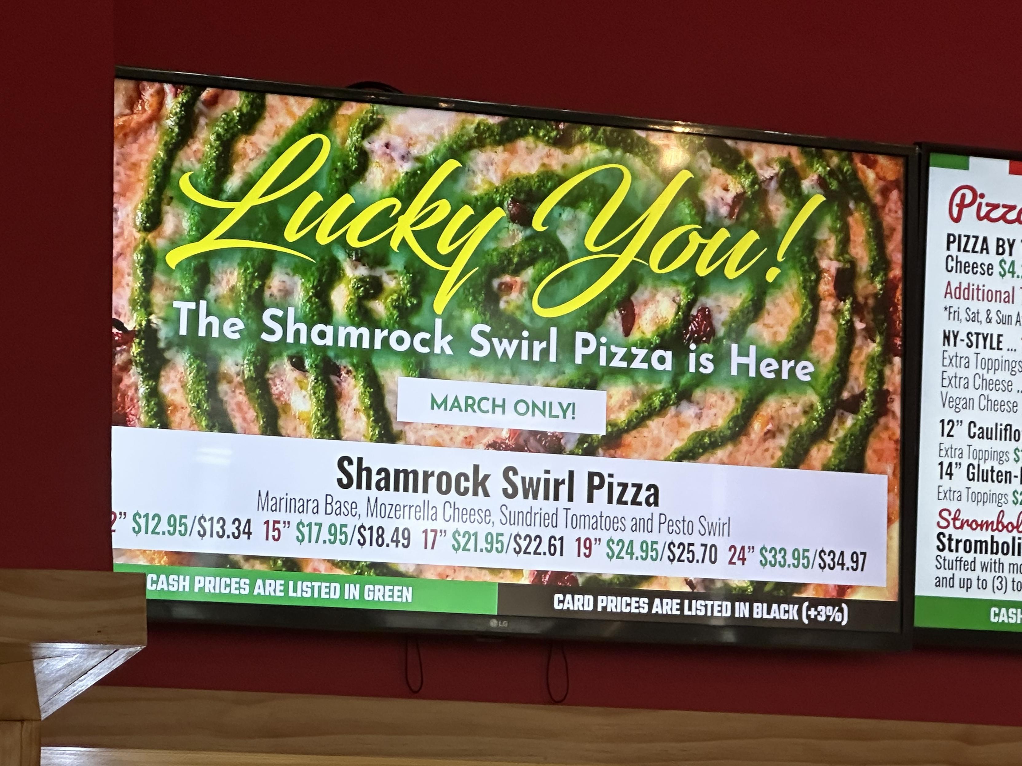 Screenshot of a digital menu at a local pizza joint showcasing a new pizza for March - Shamrock Swirl Pizza: mozz cheese, basil, sun-dried tomatoes and pesto sauce in a giant green swirl forming a spiral on the pizza.