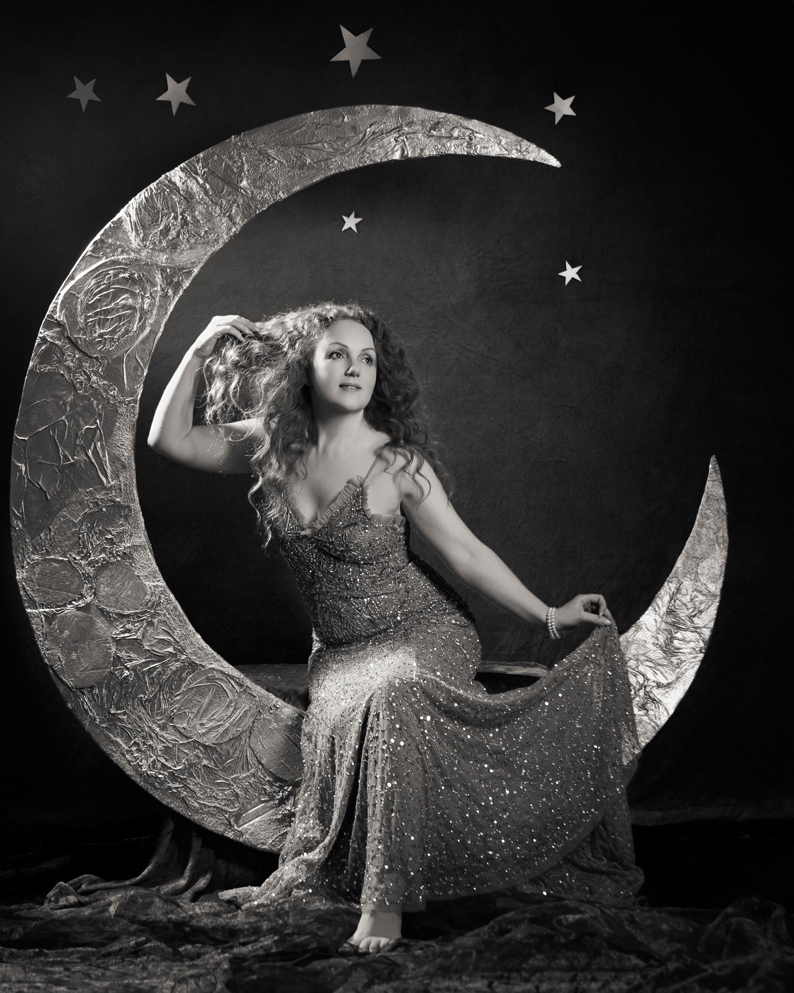 A black and white photo of model Ivory Flame, dressed in a glittery dress, sits in a large paper moon (of her own design!), teasing her hair.