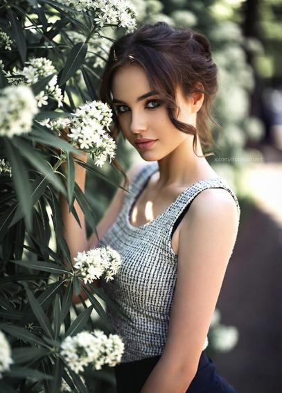 Primer plano de una mujer joven con cabello castaño recogido y ojos verdes intensos, mirando directamente a la cámara. Viste un top de tweed gris y blanco sin mangas y toca suavemente un arbusto con pequeñas flores blancas a su lado. La luz es natural y suave, creando una atmósfera tranquila.