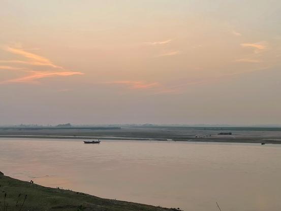 A wide, calm river landscape at dusk, viewed from a grassy bank. The sky is filled with a soft gradient of pink, peach, and grey clouds. A solitary small wooden boat with a figure is on the water in the middle ground. In the far distance, across a large sandbar, is a low, hazy treeline and distant, indistinct structures under the horizon. The entire scene is tranquil and serene.