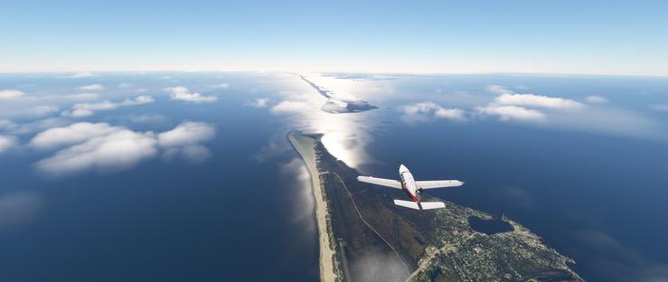 A small single-engine airplane with a white fuselage and red tail section is in flight, positioned toward the right side of the frame. It appears to be descending or approaching an airport situated on a narrow coastal landmass. The aircraft is above a light-colored sandy beach that runs parallel to the dark blue ocean, where sunlight reflects off the water’s surface, creating a bright pathway of light. The ocean extends to the horizon, with scattered white clouds floating in the clear blue sky above. To the left of the aircraft, the ocean is partially covered by low-lying clouds, while to the right, the water remains deep blue and open. The coastal landmass features a mix of dark green forested areas, brownish terrain, and a visible paved runway near the beach, with smaller clusters of buildings indicating a populated area. In the distance, small islands and rocky outcrops are visible along the coastline, adding geographic detail. The overall scene conveys a calm, sunny day with the aircraft navigating near the coastal region.