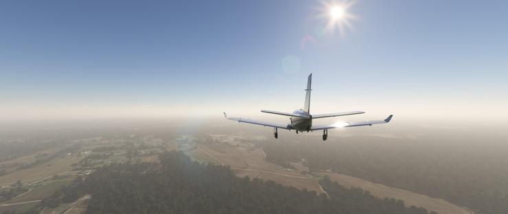 A single-engine turboprop airplane flies steadily in the sky, landing gear extended, suggesting an approach to land. The aircraft is centered in the frame, viewed from the rear, with wings level and a tail fin visible. Above, the sun shines brightly, casting a clear blue sky with subtle lens flares from the light. Below, the terrain features rolling fields in varying shades of green and brown, interspersed with dense forest areas. A faint haze hovers near the horizon, blending into the distant landscape that includes scattered buildings and winding roads. The scene depicts a serene daytime flight over a rural region, with the aircraft maintaining a stable altitude against the backdrop of open sky and earthy terrain.