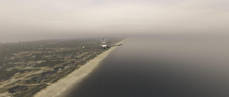 A small single-engine propeller airplane with a white fuselage and dark wings flies shortly after takeoff low over a body of water near a narrow shoreline. The aircraft is positioned just above the water's surface, moving parallel to the coast. Below, the shoreline features a sandy beach bordered by a cluster of houses with varied roof colors, including blue and dark tones, interspersed with trees and open land. Beyond the houses, a dense forest area stretches toward the horizon. The water extends into the distance, merging with a thick layer of fog that obscures the sky and creates a muted, overcast atmosphere. The fog blankets the entire scene, reducing visibility and lending a soft, grayish haze to the landscape. The airplane appears to be on a low - altitude flight path, likely approaching or departing from a nearby landing area. The overall setting is a calm, coastal environment under foggy conditions.