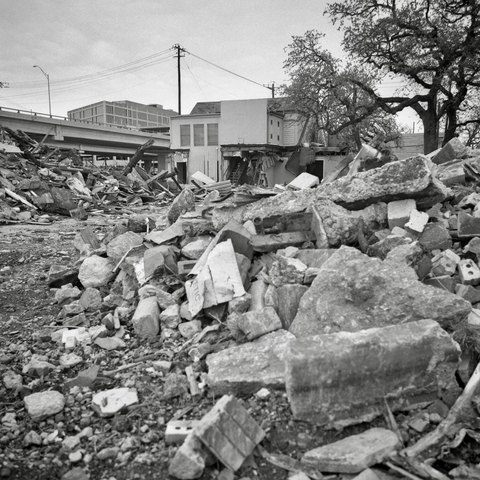 A black-and-white film photo. Large piles of debris are in front of a partially demolished building along the I-35 feeder road in East Austin, Texas. Sunday, March 8, 2026.