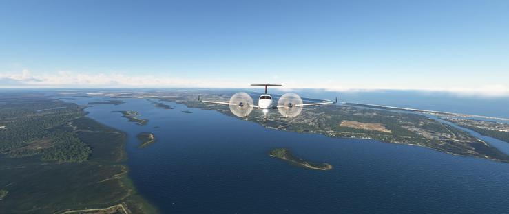 A white twin-engine propeller airplane, marked by a red tail fin, flies steadily above a vast expanse of deep blue water. Landing gear is dropped and landing lights are on, indication the plane is on final approach. Its propellers spin actively, indicating motion as it glides through the air. Below, a winding waterway cuts through a landscape of green forests, brown earthy patches, and scattered small islands, some covered in dense vegetation while others show exposed terrain. To the right, a larger landmass features clusters of urban development, winding roads, and mixed greenery. In the distance, the horizon blends into a clear blue sky with a delicate layer of wispy clouds. The scene is bright and sunlit, highlighting the contrast between the aircraft's smooth flight and the expansive natural and built environment beneath it, with the open sea stretching toward the horizon.