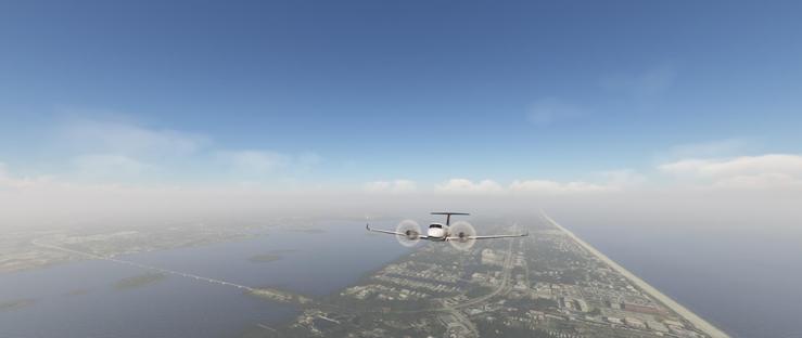 A small twin-engine propeller aircraft with a white fuselage and red accents on its tail is in flight, its propellers spinning as it glides above a coastal landscape. The plane is centered in the image, positioned above a narrow strip of land that extends into the distance, bordered by water on both sides. To the right, the coastline forms a thin, light-colored line separating the landmass from a vast expanse of deep blue water stretching toward the horizon. On the left, a larger body of water contains small islands and a bridge connecting two landmasses. Below the plane, the coastal land is densely developed with buildings, roads, and patches of greenery, while a highway runs parallel to the shore. The sky above is a clear blue with scattered wispy clouds, and a layer of low-lying haze or fog obscures distant terrain, creating a soft boundary between the sky and the land. The scene conveys a calm, clear day with good visibility, emphasizing the aircraft’s smooth flight above the expansive water and coastal development.