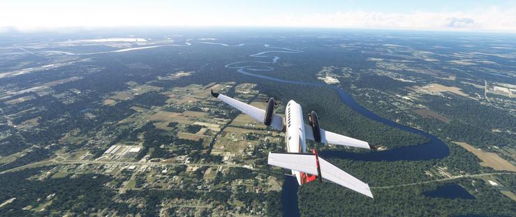 A white twin-engine aircraft with dark engines and winglets is in flight, viewed from behind. The aircraft’s tail features a red accent. Below, a winding blue river snakes through a landscape of dense green forests, brown agricultural fields, and scattered residential and industrial areas. The river meanders past clusters of houses, roads, and open farmland, while distant horizons show more terrain under a bright sky with wispy clouds. The scene captures a blend of natural and developed land under clear weather conditions.