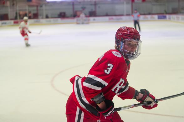 Hockey player in red clothes with white number 3 close to the front with mostly empty seeming ice behind with two other players and a ref.