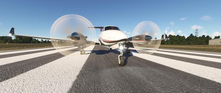 A twin-engine propeller airplane, predominantly white with a dark stripe and red accent line along the fuselage, is positioned on a paved runway. Both propellers are actively spinning, creating visible motion blur effects, and the aircraft's landing gear is deployed on the tarmac. The runway features alternating dark gray and white striped markings that extend into the distance. Beyond the runway, a flat landscape stretches out with dense green trees under a clear blue sky. To the right, a tall electricity pylon and a low-lying industrial building are visible, while the left side shows open fields and distant utility structures. The aircraft's navigation lights are illuminated, suggesting active preparation for takeoff under bright daylight conditions.
