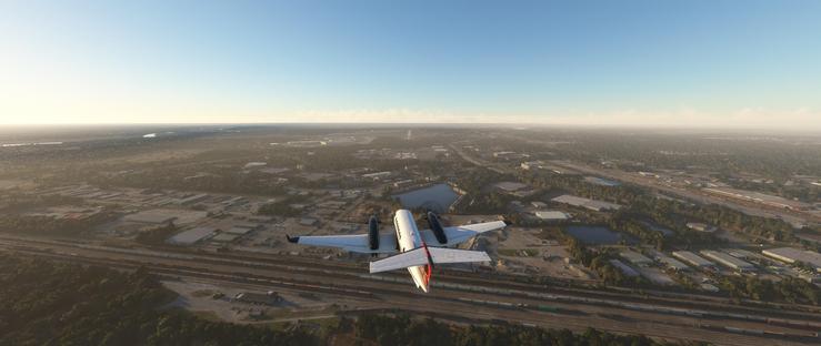 A white twin-engine jet airplane with dark engine pods and a red tail stripe is flying at low altitude, positioned centrally in the frame. It is in the process of descending for landing. Below the aircraft, multiple parallel railway tracks stretch across the scene, some carrying freight trains. Adjacent to the tracks, industrial structures—including large warehouses, low buildings, and paved lots—are interspersed with clusters of trees and small water bodies. The landscape extends into the distance, featuring more urban development, open fields, and a faint haze under a clear blue sky. The sun's position suggests early morning or late afternoon, casting a warm glow and long shadows over the terrain. The environment combines urban industrial areas with natural elements like vegetation and ponds, creating a mix of built and natural features.