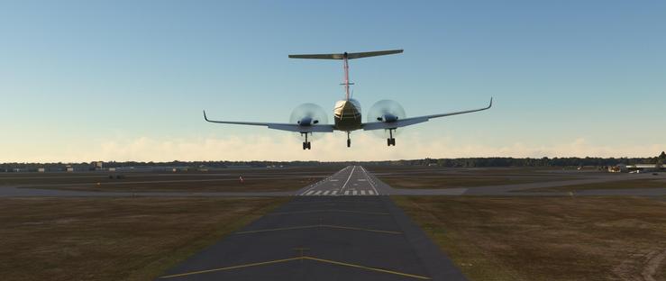 A twin-engine turboprop aircraft descends toward a runway with its landing gear extended and propellers spinning rapidly, creating subtle motion blur. The plane is centered above a paved runway marked by continuous yellow centerline dashes and crosswalk markings. Flanking the runway are expansive grassy fields that transition into a tree-lined horizon under a clear blue sky with faint, wispy clouds near the horizon. Beyond the runway, the airport features distant taxiways, small structures, and low-lying buildings, all illuminated by bright daylight. The scene captures a smooth landing approach with excellent visibility, calm weather, and minimal turbulence, emphasizing the aircraft’s steady descent toward the paved surface.
