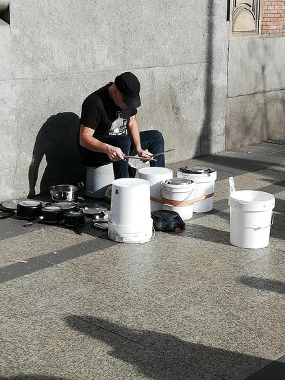Auf dem Gehweg sitzt ein Mann mitten zwischen umgedrehten Farbeimern und Edelstahltöpfen und trommelt darauf. Vorne steht ein grosser Eimer für die Spenden.