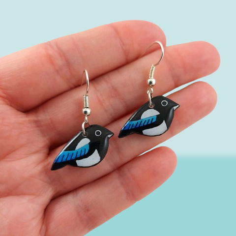 Boucles d'oreilles fantaisie pendantes éco-responsables en forme de petites pies bleues.
Les perles sont réalisées en CD recyclé, découpées et peintes à la main, puis recouvertes de résine.
Les attaches sont en laiton argenté, mais sont aussi disponibles dans d'autres métaux et coloris.
Ces boucles d'oreilles sont tenues entre les doigts tendus d'une main ouverte devant un fond bleu pastel.