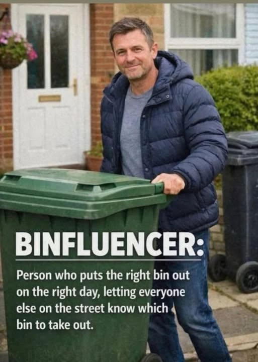 A white male dressed in dark winter coat, t-shirt and jeans, smiling, while he is steering a large green garbage bin past a black one, as he walks it to the curb.
Caption : Binfluencer: Person who puts the right bin out on the right day letting everyone know which bin to take out.