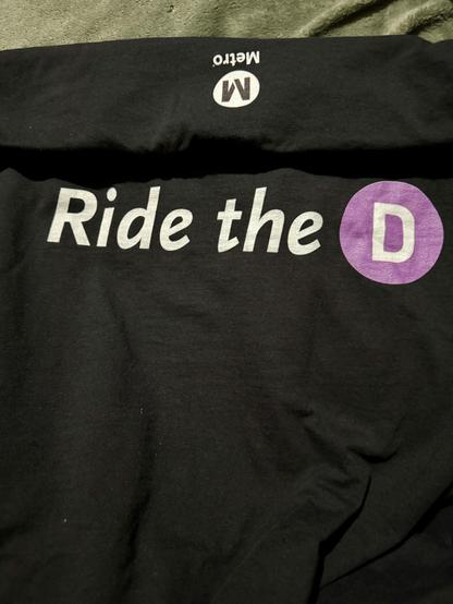 Black T-shirt that has the LA Metro logo on the back and the front says Ride the D. The D is in white in a purple circle representing the D line on LA Metro.