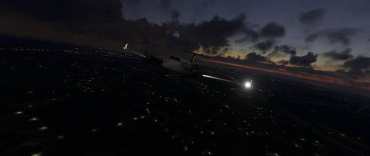 A small white turboprop aircraft flies low over an urban area during twilight. Its landing lights illuminate the dark terrain below, which is dotted with numerous city lights from buildings and streets. The sky above is darkening, with heavy clouds partially obscuring a faint orange and red sunset near the horizon. The aircraft's nose is slightly angled upward, and its wings extend outward with visible engine nacelles. Below, the landscape shows a mix of illuminated urban zones and darker rural areas, with faint light trails suggesting highways or rail lines. The overall scene conveys nighttime flight with a dramatic sky contrasting the artificial lights of the city and the natural twilight hues.