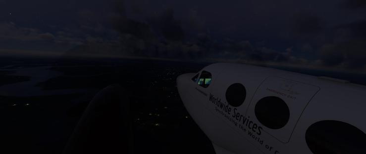 A white commercial airplane is in flight during nighttime, viewed from outside. The aircraft’s nose and cockpit window are prominent, with an interior screen visible through the glass. The plane’s fuselage displays printed text including “Worldwide Services” and “Synchronizing the World of C(ommerce)” along with an “EMERGENCY EXIT” label. The aircraft is moving above a dark landscape where scattered city lights illuminate urban areas, with a vast expanse of water visible in the distance under a starry sky filled with dark clouds. The scene depicts the plane traveling over land and ocean at night, with the horizon line separating the illuminated city below from the night sky above. The sky shows a mix of deep blues and darker cloud formations, while the ground features patches of light from populated regions and darker, undeveloped areas.