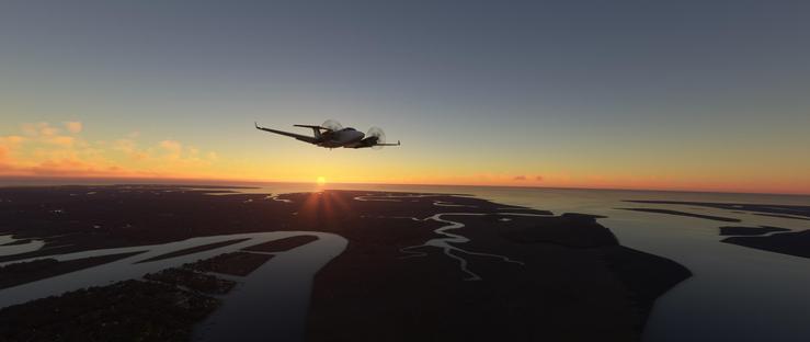 A small twin-engine propeller aircraft flies low over a coastal landscape during sunrise, its silver fuselage and wings visible against the sky with both propellers spinning rapidly. Below, a vast plain stretches toward the ocean, marked by intricate, winding waterways that cut through dark terrain. The horizon reveals a distant sun setting, emitting vibrant orange and pink light that blends into a deep blue sky. Wispy clouds near the horizon catch the sunlight, creating a soft glow. The land features patchwork fields and dense clusters of trees, while the waterways reflect the sunset’s warm hues. In the distance, the ocean meets the sky, forming a seamless line where the sun dips below the horizon. The aircraft’s position and altitude suggest it’s navigating over the intricate network of rivers and estuaries.