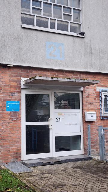 A door with large glass pane, and a white plastic frame, and four letterboxes integrated. The building has exposed red bricks, and a blue sign is attached to the left of the entrance.