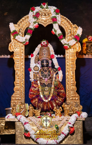 Idol of Hindu deity Krishna in gray (broad red border), standing on a gold covered pedestal, holding a staff (not fully visible) in right hand, left hand on front left waist; wearing gold shoes gold palm coverings, a stone-studded crown, and lots of gold ornaments. A string of white flowers (some red roses) around Krishna waist-to-waist. An ornate gold arch behind Krishna. A string of white flowers (some red bands) hooks from the left side of the arch to the middle and from there to the right. Some smaller idols on the pedestal. A string of white flowers (some red roses) on the pedestal. Dark blue cloth background.