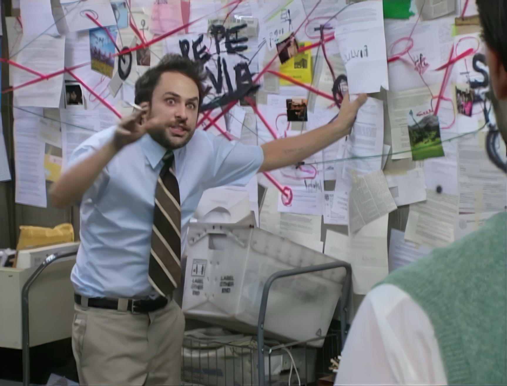 Screenshot from the show it's always sunny. A guy in a button-down short sleeve shirt and tie stands in front of a wall, covered with documents and red string connecting documents. He's wide eyed and smoking a cigarette.