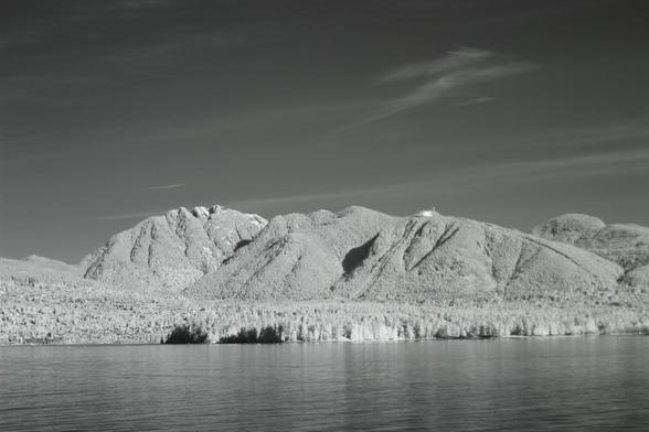 Very clear image of woods-covered mountains on the other side of a lake with a few long clouds in the background under a mostly dark sky