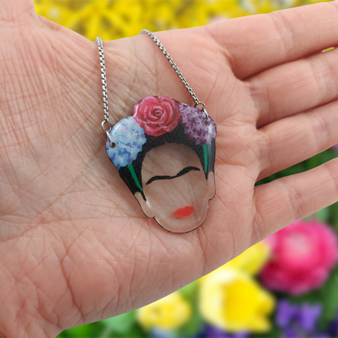 Collier avec pendentif éco-responsable à l'image de Frida Kahlo coiffée de 3 fleurs (personnalisables).
Ici, il s'agit de fleurs d'hortensia bleue, d'une rose rouge, et de lilas violet.
La perle est réalisée en CD recyclé, découpée et peinte à la main, puis recouverte de résine.
La chaine réglable est en acier inoxydable argenté ou doré.
Le collier est tenu dans le creux d'une main ouverte. En arrière-plan, un parterre de fleurs jaunes, rose et violette.