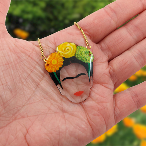 Collier avec pendentif éco-responsable à l'image de Frida Kahlo coiffée de 3 fleurs (personnalisables).
Ici, il s'agit de fleurs de gerbera orange, d'une rose jaune, et de chrysanthème vert.
La perle est réalisée en CD recyclé, découpée et peinte à la main, puis recouverte de résine.
La chaine réglable est en acier inoxydable argenté ou doré.
Le collier est tenu dans le creux d'une main ouverte. En arrière-plan, un parterre de fleurs orange.