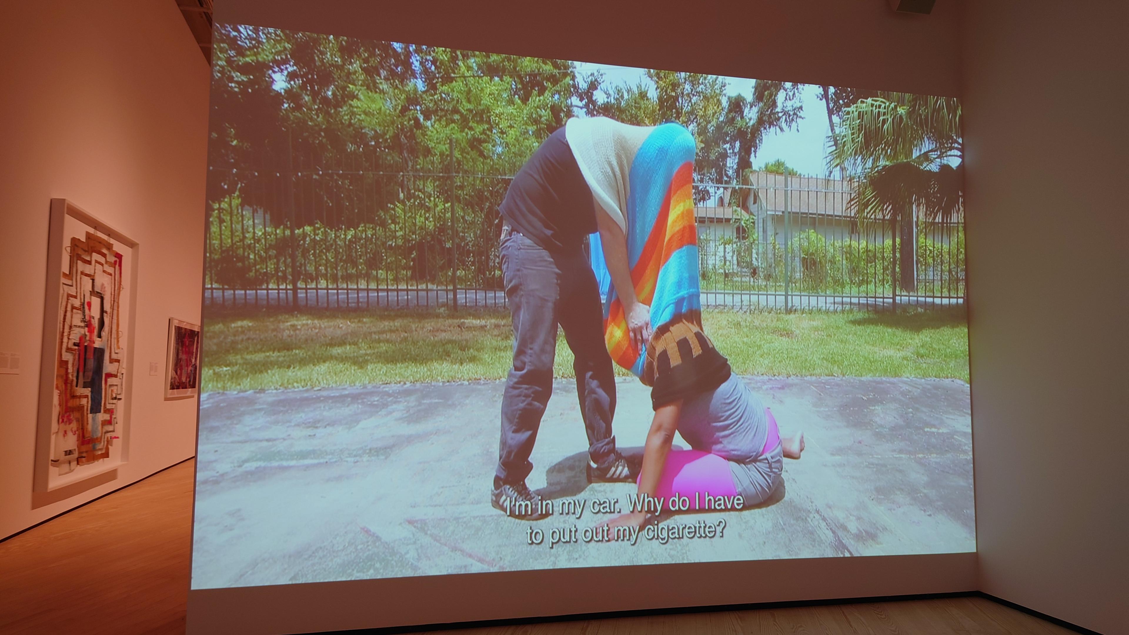 Art gallery. On one wall two people with their head stuck in the same sweater are tangled on pavement. Caption: "I'm in my car. Why do I have to put out my cigarette?"