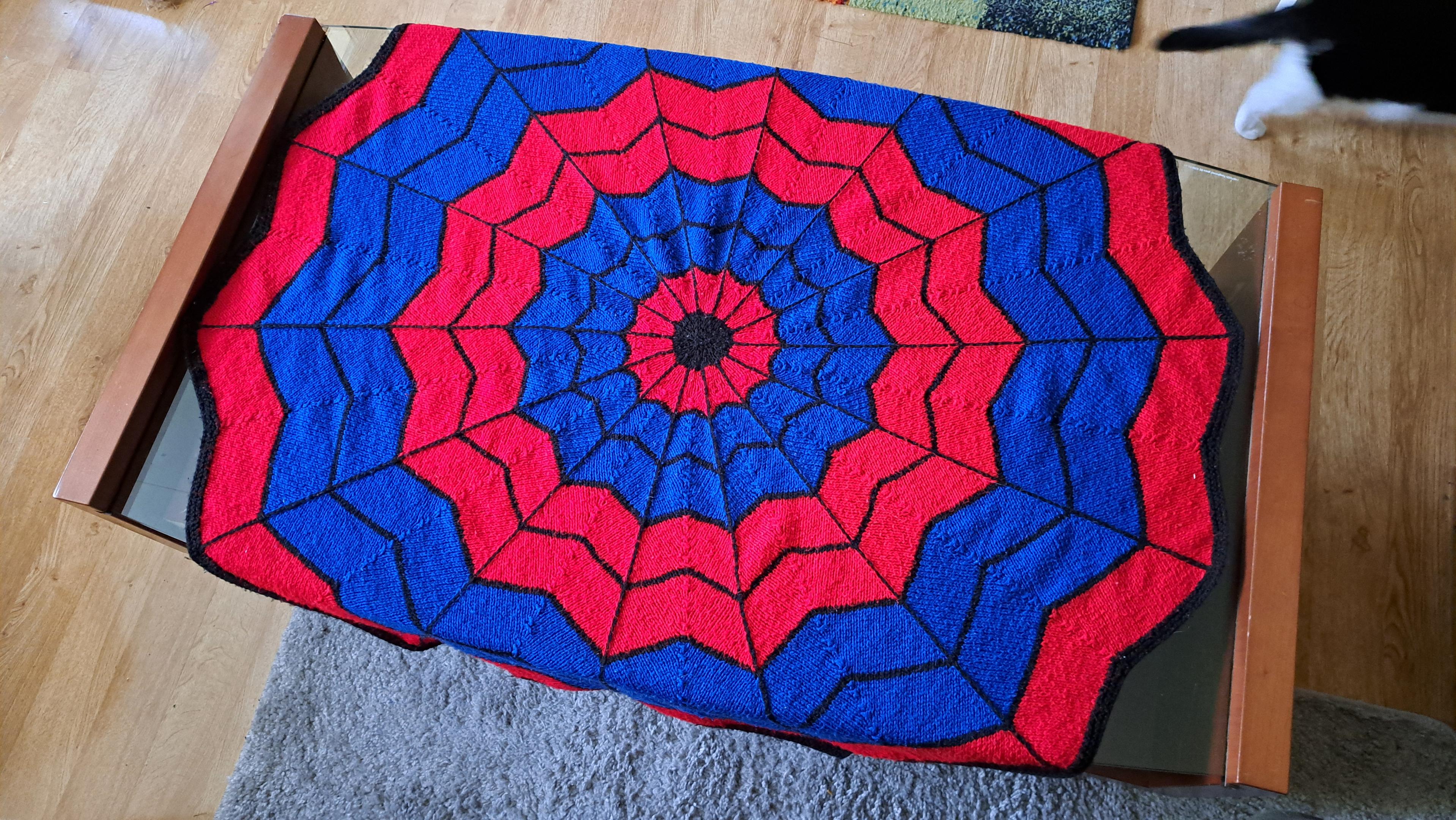 A circular, web-shaped blanket, made of alternating red and blue circles with a web design, is laid out over a large glass coffee table which it fits perfectly, almost as though it were designed to be put there.
