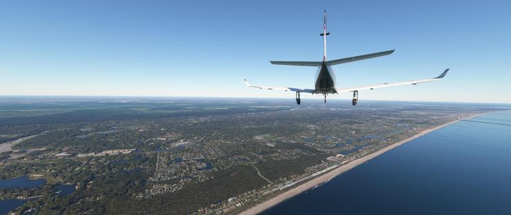 A small airplane with a white fuselage, dark tail, and extended landing gear is descending toward a coastal area. Below, a long sandy beach curves along the edge of a deep blue ocean, bordered by dense green forests and scattered residential neighborhoods. The landscape features numerous small lakes and winding waterways, with flat terrain extending into the distance under a clear, bright blue sky. The aircraft is positioned centrally above the coastline, with its landing gear lowered, indicating preparation for landing near the populated shoreline. The scene captures a smooth approach to land over a mix of natural and developed areas, with the ocean on one side and the horizon visible under a cloudless sky. The terrain below includes patches of developed land, forested regions, and small bodies of water, all under a vast expanse of clear blue sky.
