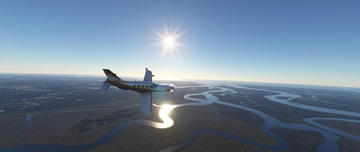 A small twin-engine private jet with a white fuselage, dark blue tail, and red accent stripe is in flight, positioned slightly left of center. The aircraft flies low, its nose angled slightly upward, suggesting steady navigation. It moves across a vast landscape defined by a complex network of winding rivers that reflect sunlight into a bright, shimmering ribbon. The sky is clear and deep blue, with the sun high in the sky creating a pronounced lens flare. Below, the terrain is predominantly brown and arid, interspersed with clusters of green vegetation, while distant horizons blend into a hazy line. The plane’s tail displays the letters TMC in dark text against the light background. The scene depicts a serene aerial journey over a delta-like region under a bright, clear day.