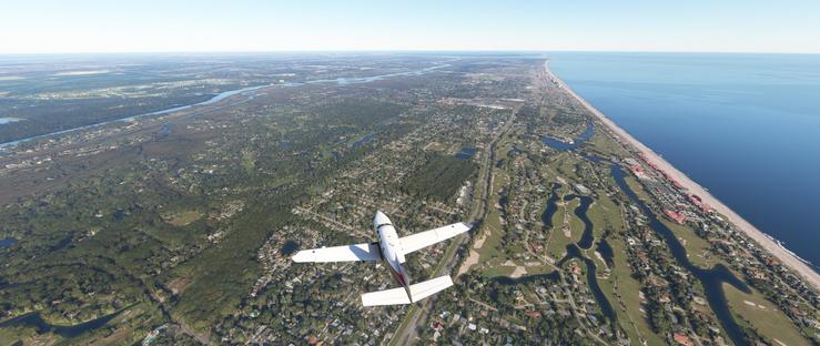 A small white single-engine airplane flies low over a coastal landscape. Below, a sandy beach curves along the ocean’s edge, bordered by a strip of residential neighborhoods with red-tiled roofs. To the left, dense green forests and scattered waterways, including a winding river, stretch toward the horizon. Interconnected roads and patches of developed land with houses and parks are visible. In the distance, the ocean meets the sky under a clear blue atmosphere. The scene captures a sunny day with the plane positioned centrally, moving over a mix of natural and human-made environments.