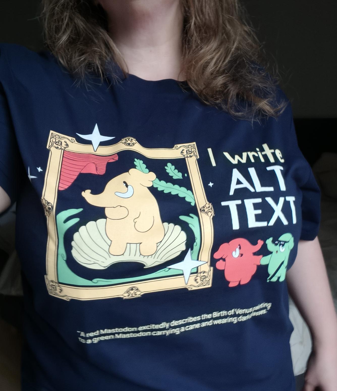 Tragefoto vom neuen Mastodon-Tshirt.
Dunkelblaues T-Shirt mit Aufdruck
Eine große gelbe Mastodon-Figur befindet sich als Venus-Gemälde in einer Muschel, die von einem goldenen Rahmen umgeben ist. Rechts daneben stehen zwei kleine Mastodons wie im Museum vor dem großen Gemälde, die grüne Mastodon-Figur mit einem Stock und dunkler Brille, ein rotes Mastodon erklärt ihm das Bild. Oben rechts neben dem Gemälde steht der Text 'I write ALT TEXT'.
Unten ist ein kleiner weißer Text: 'A red Mastodon excitedly describes the Birth of Venus painting to a green Mastodon carrying a cane and wearing dark glasses.'