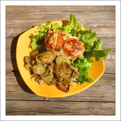 A bright yellow, slightly asymmetrical plate sits on weathered wooden boards, catching warm sunlight that throws crisp shadows across the scene. On the plate a bed of fresh, ruffled green lettuce forms a lively backdrop for a rustic, homey meal. Two halved tomatoes, their cut faces topped with bubbling, golden-browned cheese, perch near the top — the cheese has browned in spots and the tomato edges are slightly roasted, giving them an inviting, savory gloss.
Below and spilling toward the front of the plate is a generous heap of thinly sliced potatoes, browned at the edges and mingled with translucent, caramelized onions. The potato slices look tender and layered, with some pieces curling and crisping where they've browned, suggesting a comforting mix of soft interior and crisp exterior. The overall color palette — vivid green, rich red, warm golden-browns on the food set against the sunny yellow plate and rustic wood — feels warm and appetizing, as if the dish has just been set out for a casual, sunshine-lit meal.