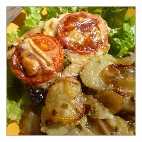 A close, mouth-watering shot of a rustic plate of roasted vegetables. In the foreground are thin, golden-brown potato slices — their edges are crisped and slightly caramelized, flecked with chopped herbs (looks like chives or parsley) and translucent ribbons of onion. Two small tomato "cups" sit atop the potatoes: each tomato half has been roasted or baked until the skin darkens and the surface bubbles with melted, browned cheese, creating little glossy, savory crowns. Bright green lettuce leaves form a fresh backdrop, their ruffled texture contrasting with the warm, glossy vegetables. The overall color palette — warm ambers and golds against leafy green — and the close framing make the dish look both homey and appetizing, with visible textures that suggest crispness, softness, and a tangy, savory finish.