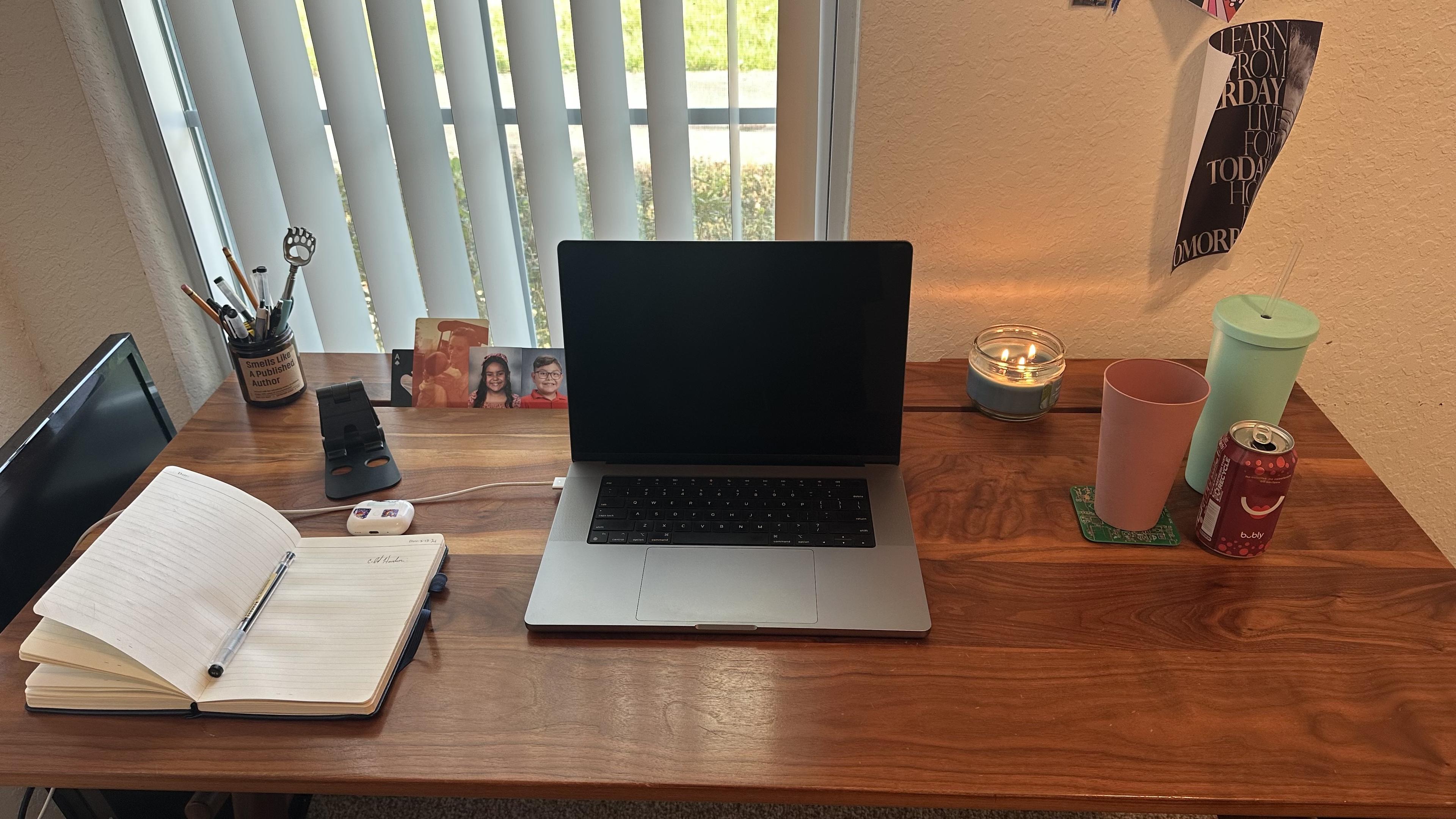 My desk. Laptop in the middle. To right: a cup, can of sparkling water and cup with straw. To left, notebook and pen, cup of pens.