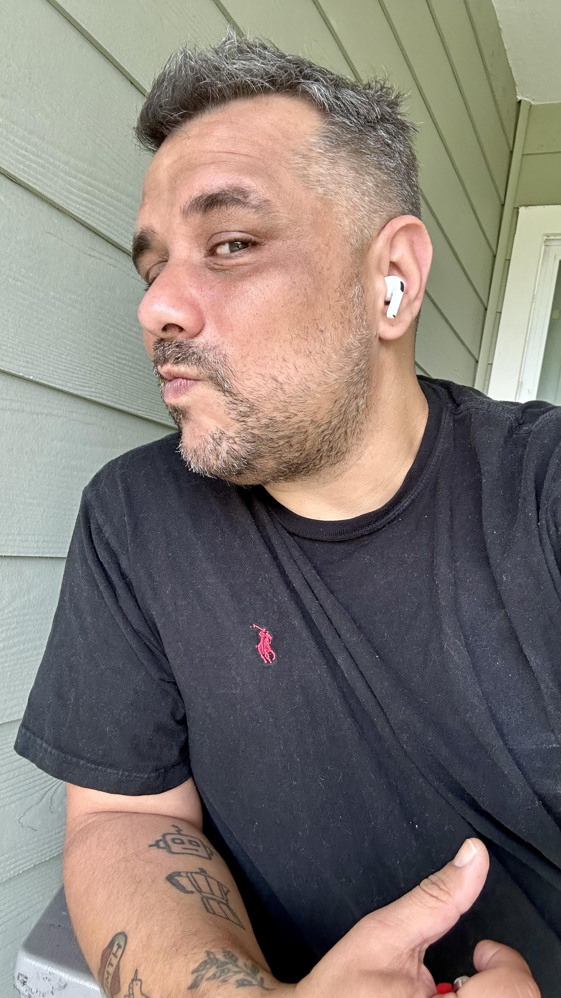Me side profile showing off my hair cut. I got earbuds in. Wearing a black t-shirt. Sitting outside on the patio.