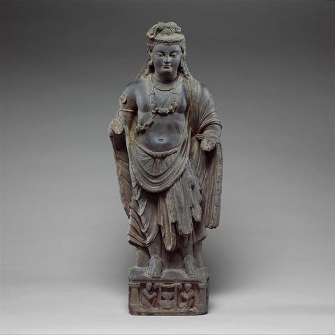 a statue , it is dark colored, it is a buddha standing wearing jewels and fine heavenly clothing, looking downwards, it is very lifelike and beautiful , the expression very peaceful on it