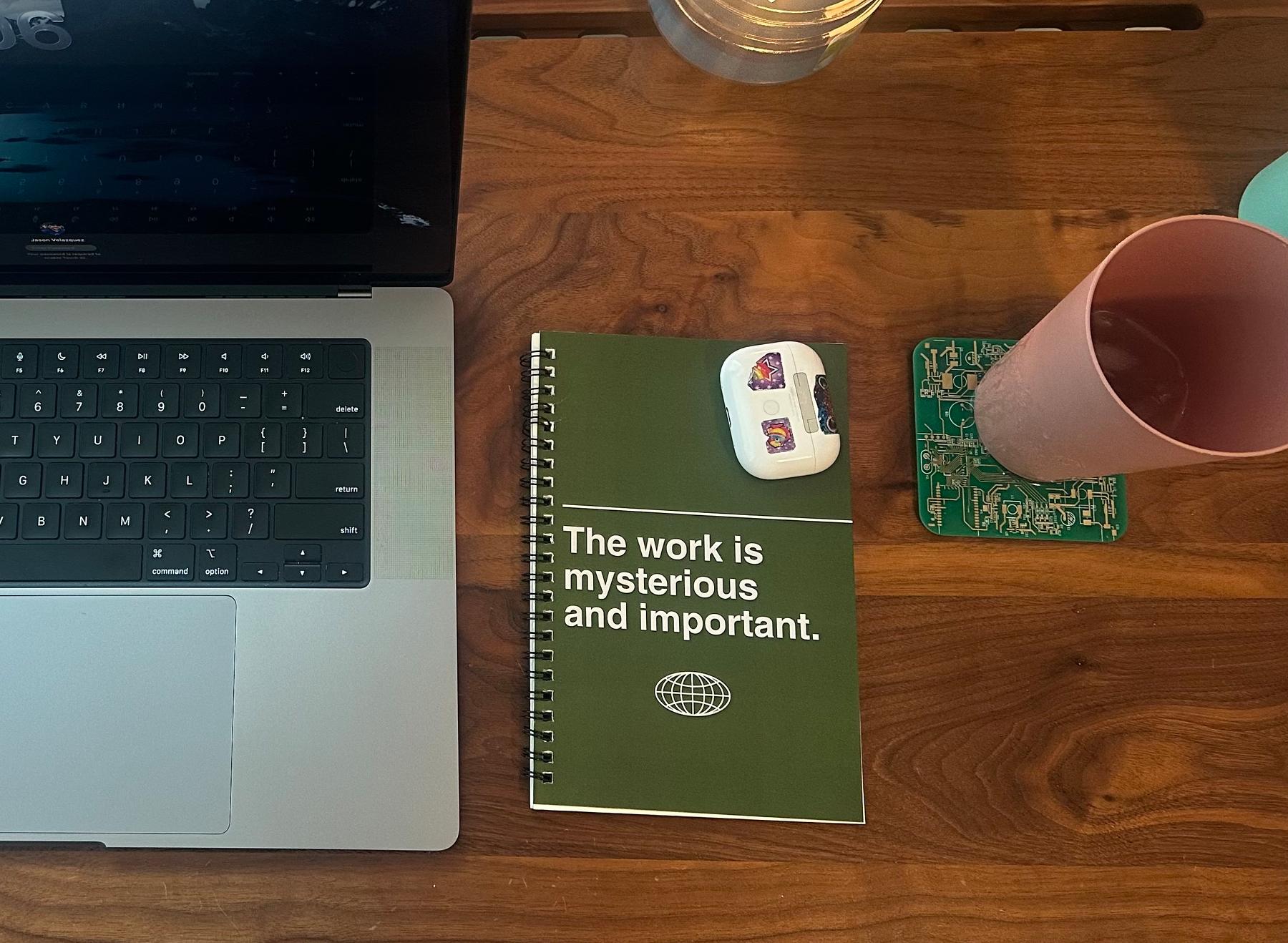 Over top shot of my desk. Laptop, earbuds, a cup and a notebook that reads "the work is mysterious and important."