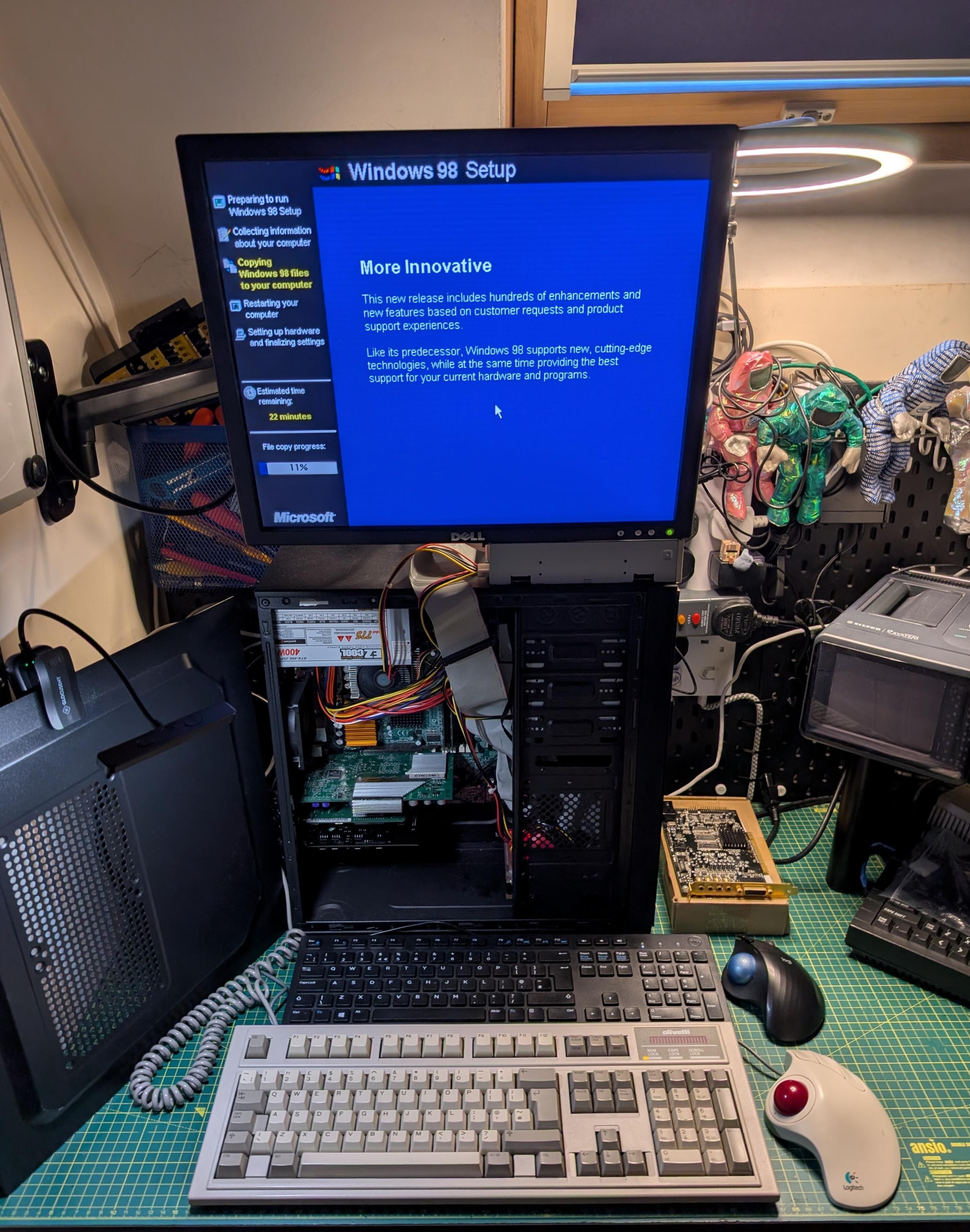 A retro computer setup on a workbench with an open desktop PC case running the Windows 98 Setup screen on a Dell monitor. The installer shows file copying progress at 11%. Inside the case, IDE ribbon cables and components are visible. In front of the system are two vintage-style keyboards, including an Olivetti keyboard, along with a trackball mouse. The desk is covered with a green cutting mat and surrounded by electronics tools and vintage equipment, including a portable CRT TV and PCI Soundcard, giving the space a retro computing workshop feel.