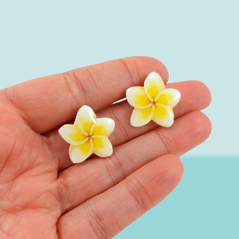 Puces d'oreilles éco-responsables en forme de fleurs de frangipanier blanches et jaunes, réalisées en CD recyclé et peintes à la main.
Elles sont tenues entre les doigts tendu d'une main ouverte devant un fond bleu pastel.
Les tiges sont en acier inoxydables.