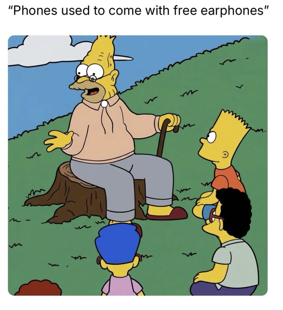 A meme featuring Grandpa Simpson from "The Simpsons" sitting on a tree stump, gesturing as he tells a story to Bart, Milhouse, and Nelson. The text at the top of the image reads, "Phones used to come with free earphones".